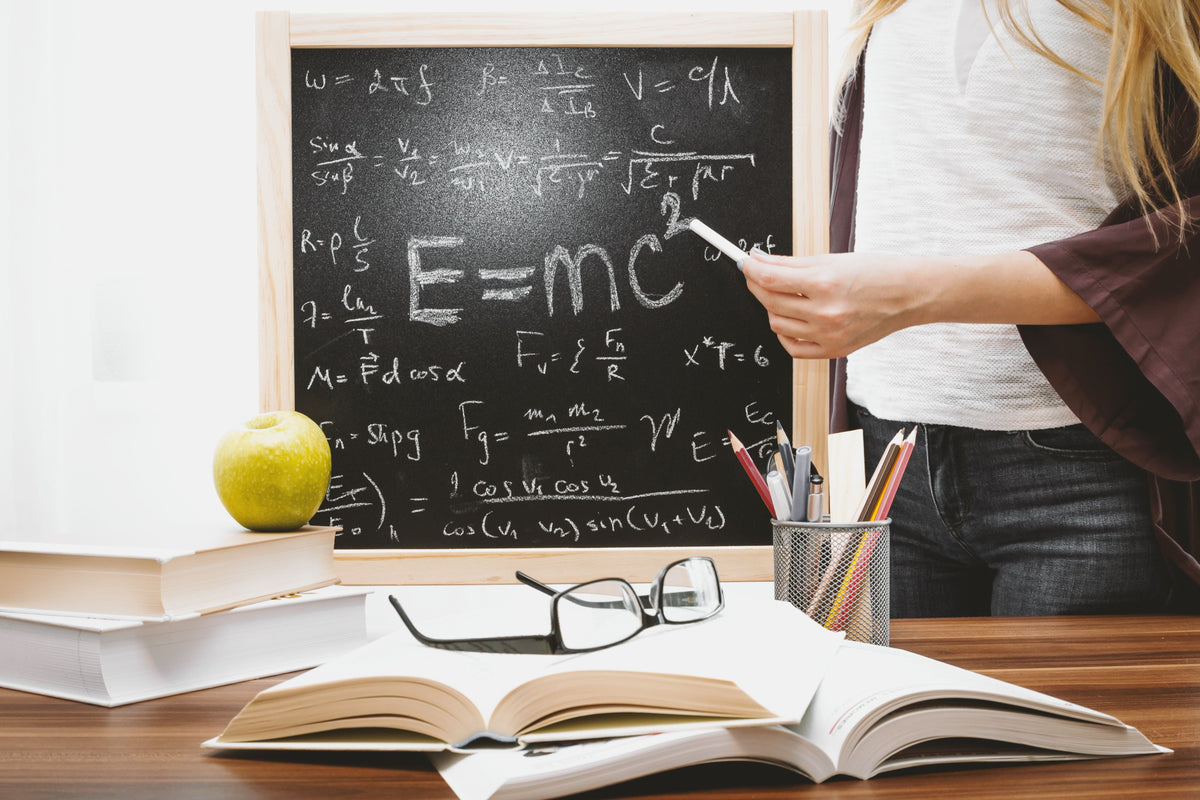 Woman writing physics equations on a blackboard with books and an apple on the desk. Image by Jeshoots.com@pexels.com